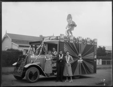 Image: Unidentified men and women in front of a truck advertising Krane Mild Cheese, with an elfin cutout holding a cheese box on top of the truck, and cheese box packets stringed together in front of the windscreen, location unidentified
