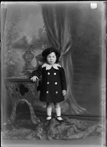 Image: Studio portrait of unidentified young girl in a dark hat and double breasted coat with a large lace collar standing next to a wooden high chair, Christchurch