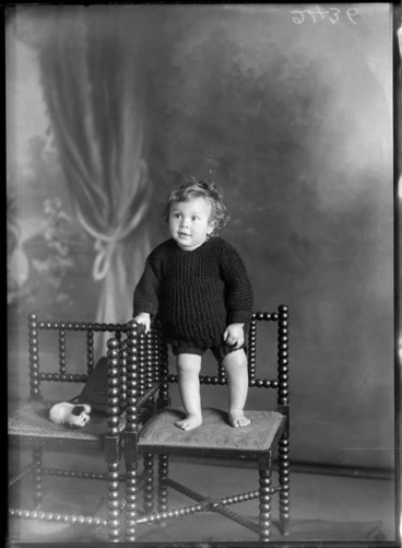 Image: Studio portrait of an unidentified boy, dressed in a two piece woollen jumper suit with shorts, standing on a wooden seat, with a fluffy stuffed toy dog and handbag on a seat next to him, possibly Christchurch district