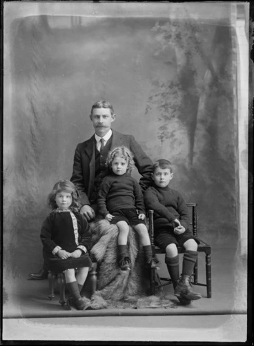 Image: Studio portrait of members of an unidentified family, a man, two boys with a girl, showing the girl sitting on a wooden stool, dressed in a dark velour dress, with patterned borders on the dress, with a ribbon in her hair, the young boy sitting on a fur covered box in front of the man, dressed in a high neck jersey, with velour shorts, with mid length socks, dark leather shoes with large laces, with the other boy sitting on a wooden chair, dressed in a high neck woollen jersey, with velour shorts, knee high woollen socks with stripes, with ankle laced boots, possibly Christchurch district