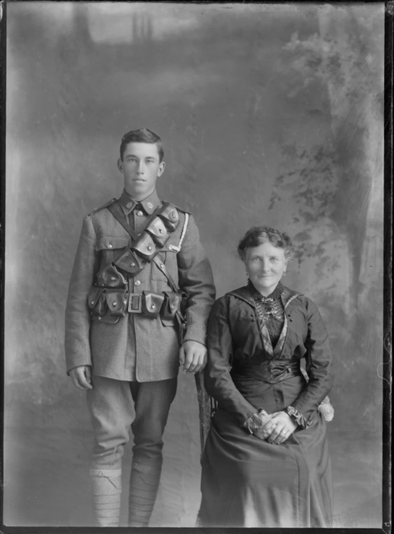 Image: Studio unidentified family portrait of older mother sitting in a dark lace blouse and dress next to her young adult son in World War I soldier's uniform with bandolier and waist ammunition belts, collar badges and white shoulder pocket cord standing, Christchurch