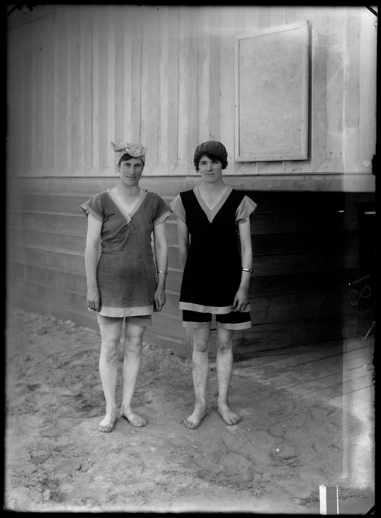 Image: Two unidentified women, dressed in bathing suits and wearing swim caps, outside a shed at the beach, probably Christchurch district