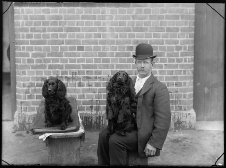 Image: Unidentified man, outside a brick building, with two dogs, one is sitting on his lap, probably Christchurch district