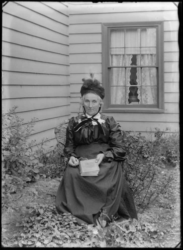 Image: Unidentified elderly woman, sitting outside a house in a garden, holding a book and spectacles, probably Christchurch district