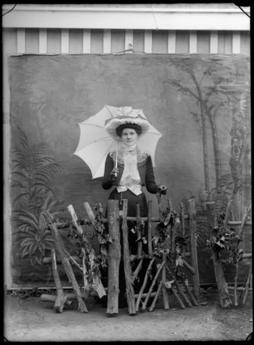 Image: Portrait of unidentified woman, behind a rusticated prop fence, holding an opened parasol, with a painted studio backdrop, probably Christchurch district