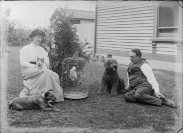 Image: Unidentified man and woman with domestic pets, including two dogs, a cat, and a bird in a cage, sitting on a grass lawn outside a wooden building, possibly Christchurch district