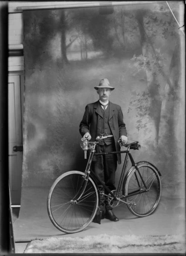 Image: Studio unidentified portrait of an older man with large moustache, three piece suit and hat, standing with a men's bicycle with carbide headlamp, Christchurch