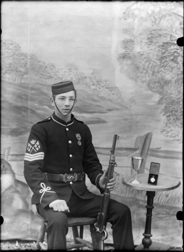Image: Outdoors in front of false backdrop, a young World War I sergeant in dress uniform with lapel medal, flag and crown sleeve badges and insignia, sitting with a rifle and table with small cup and medal, probably Christchurch region