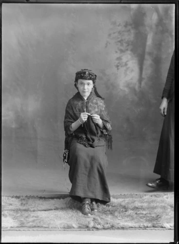 Image: Studio portrait of unidentified girl dressed as an elderly woman, with glasses, shawl, dark floral hat and knitting a scarf, Christchurch