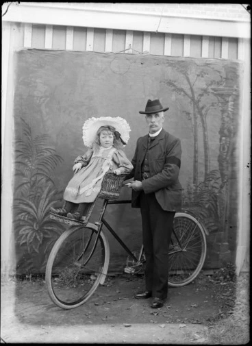 Image: Outdoors portrait of man holding a bicycle and a young girl with large hat is on a front handlebars wicker child carrier, probably Christchurch region