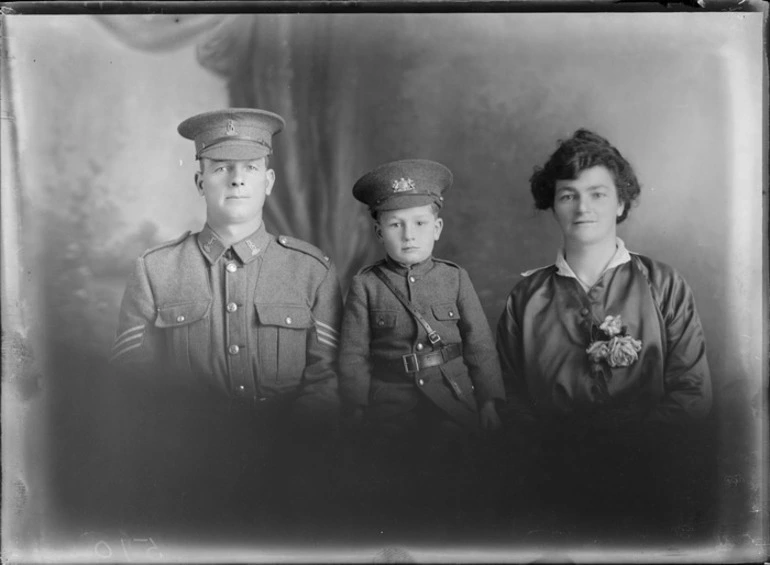 Image: Studio upper torso unidentified family portrait, a Sergeant World War I soldier with collar and hat badges sitting with young son in soldier's uniform with hat and badge, and wife with flowers, Christchurch