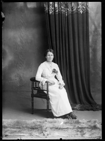 Image: Studio portrait of unidentified young woman with large square lace collar over a cotton dress and lapel corsage, with jewelled neck bar brooch, necklace and wrist watch sitting in a wooden chair, Christchurch