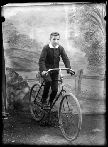 Image: Outdoors portrait of unidentified boy in an Arundel shirt collar, with tweed jacket and shorts, knee high socks and cap riding a bicycle, taken in front of false backdrop, probably Christchurch region