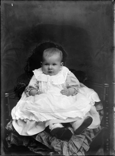 Image: Studio portrait of unidentified baby in a lace dress with large collar and booties, sitting on cushions on a wooden chair, Christchurch