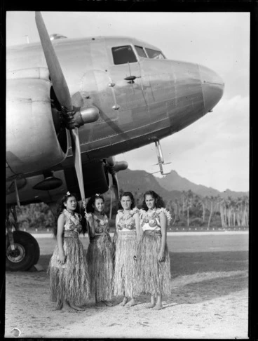 Image: Four unidentified local girls in hula skirts in front of a C47 transport aircraft, Rarotonga airfield, Cook Islands