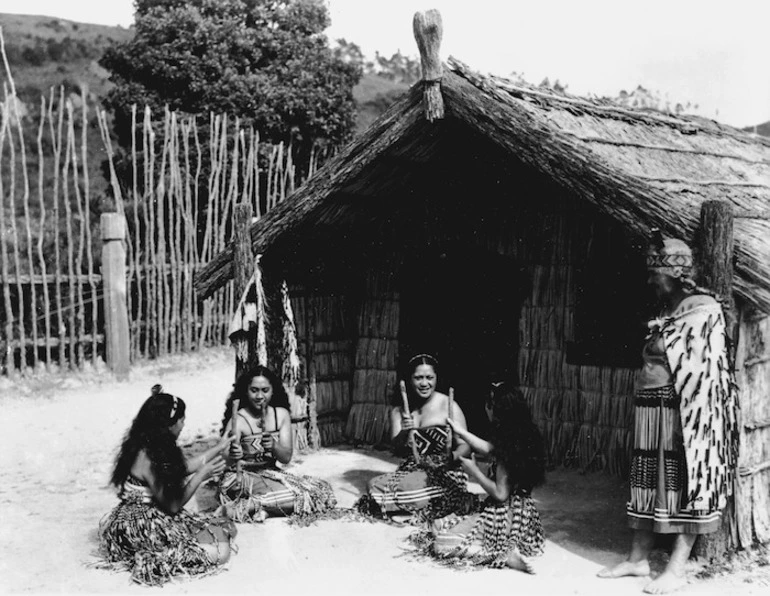 Image: Maori women, in semi traditional costume, playing a stick game alongside a meeting house