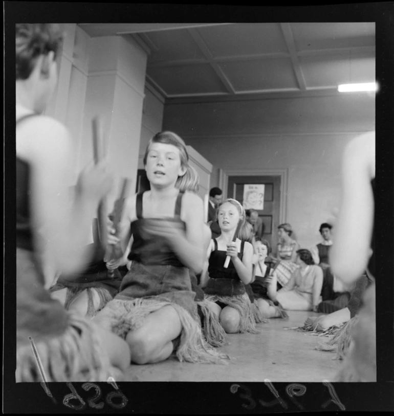 Image: Children in grass skirts kneeling and playing Maori stick games in a classroom, probably in Wellington