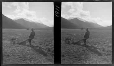 Image: An unidentified man with plough in a field, haystack in distance and mountains beyond, probably West Coast Region