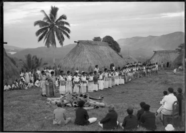 Image: Fijian women dancing, with dwellings behind, a group in the foreground watching, and another group behind the women