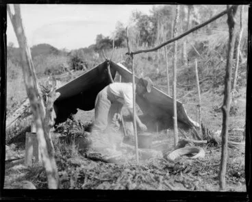 Image: Unidentified Maori man cooking over an open fire beside a tent fly; camping near the Whanganui river