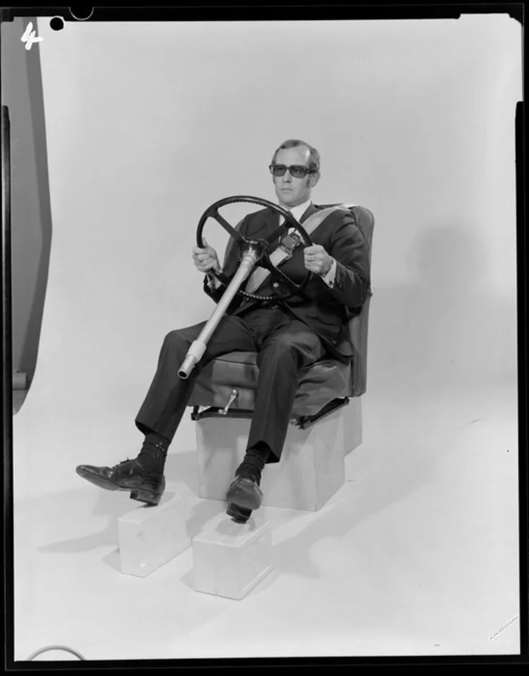 Image: Man in studio sitting in car seat, with steering wheel and seatbelt