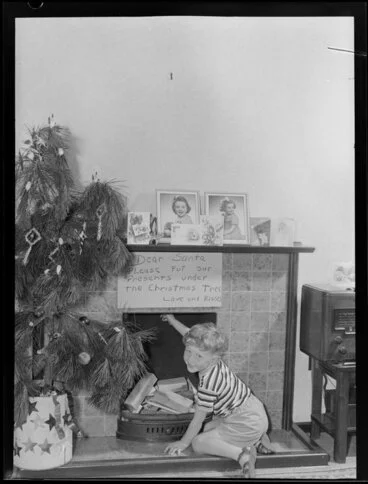 Image: Preparing for Christmas, boy beside Christmas tree with note for Santa above fireplace