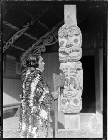 Image: Tarihira Mihinui dressed in a feather cloak holding a tewhatewha, next to a carved wooden pou, Otūkou marae