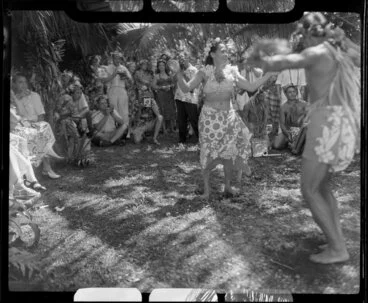 Image: Unidentified local couple, possibly including Augustine, perform the hula while tourists take photographs at a ceremony feast, Tahiti; local drummers in the background