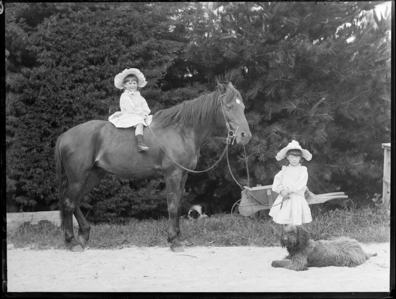 Image: Two unidentified young girls, one of whom is sitting on the horse that the other is holding, including two dogs