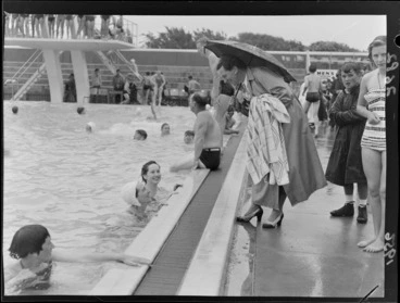 Image: An unidentified woman, sheltering under an umbrella and talking to swimmers, Naenae Olympic Swimming Pool, Lower Hutt