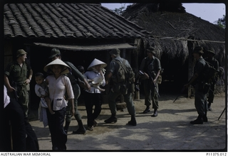 Members of B Company, 2RAR/NZ, and a Vietnamese National Policeman (partly obscured, left) chivvy women carrying children from their homes as they conduct a cordon and search of the village of Hoa ..