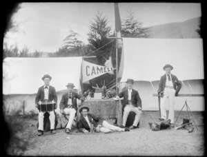 Group of unidentified men outside two tents, flag reads 'Camelia', includes man holding an accordian and other sit at a table, probably Christchurch district
