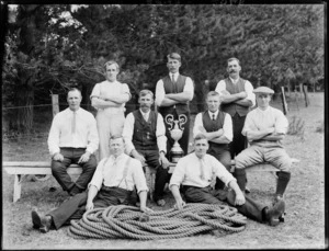Unidentified group portrait of men in shirts with an ornate cup and a coil of hawser laid rope [tug-of-war team?], pine trees beyond, probably Christchurch region