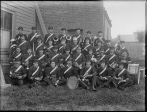 Large unidentified brass band outside a brick building, showing members in uniform with instruments, probably Christchurch district