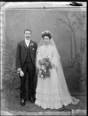 Outdoors portrait of unidentified wedding couple standing in front of wooden wall with false backdrop, bride with long veil and holding flowers, probably Christchurch region