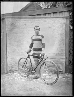 Unidentified man in cycling attire standing with his road racing bicycle within a backyard area with wooden fence behind, probably Christchurch region