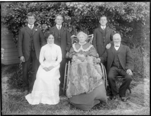 Family portrait, unidentified elderly father with mother in a wheelchair and their adult three sons and daughter, in front of a tree and house, probably Christchurch region