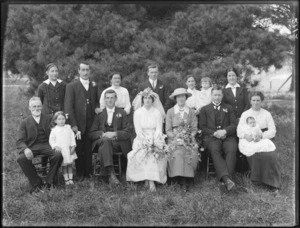 Wedding group portrait, unidentified bride and groom with adult extended family including a baby and young children, on long grass in front of pine tree, probably Christchurch region