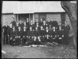 Group portrait of unidentified young men, some with handkerchiefs in breast pockets, in front of a wooden building [at camp?], probably Christchurch region