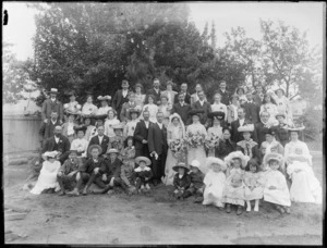 Large wedding group portrait in backyard by a fence, unidentified bride and groom with extended family with children in front, women wearing hats, probably Christchurch region