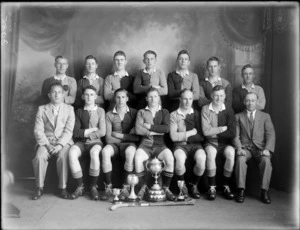 Studio portrait of men's hockey team, unidentified players and coaches with three small cups and a large trophy cup with lid and small shield plaques around its base, Christchurch