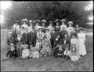Wedding party portrait, unidentified bride and groom with extended family, children in front, in a field with trees behind, women wearing hats with flowers, two older women with face veils, probably Christchurch region