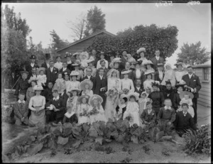 Large wedding party portrait behind vegetable garden with ivy bush, garage and house behind, unidentified bride and groom with family and friends, children and babies, women in hats, some women with face veils, probably Christchurch region