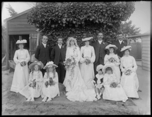 Wedding party portrait in backyard with garage and house beyond, in front of ivy bush with woman's head with hat looking down, unidentified bride with long veil and train, groom and family, four flower girls and page boy, probably Christchurch region