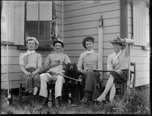 Four unidentified young men wearing hats, sitting with dog in chairs outside a wooden building with three shotguns and three baby rabbits, drinking from beer bottles at their feet, dead rabbits hanging behind, probably Christchurch region