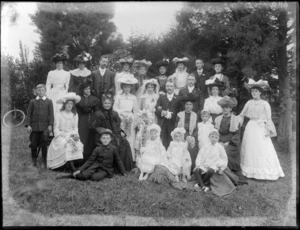 Wedding group portrait in backyard with trees and wooden fence beyond, unidentified bride and groom with extended family, flower girls and page boys, women wearing hats, older women with lace collars, probably Christchurch region