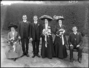 Wedding party portrait on tennis court with hedge behind, unidentified bride and groom with bridesmaids and best men, women with hats and flowers, probably Christchurch region