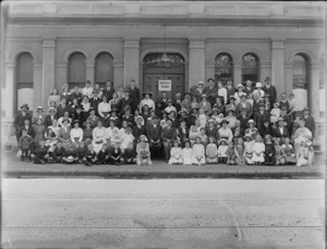 Large group portrait of unidentified men, women and children, with Bruce's Picnic' sign outside the Orange Hall building of the Loyal Orange Institution charitable organisation, probably Christchurch