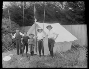 Five unidentified young boys in hats and caps, holding bamboo poles, standing in front of a tent with a flag, wooden fence and trees beyond, [Sumner?], Christchurch