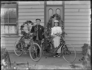 Young unidentified man with a lapel medal and women standing in front of a wooden house with ivy decorated bicycles and wreaths, probably Christchurch region